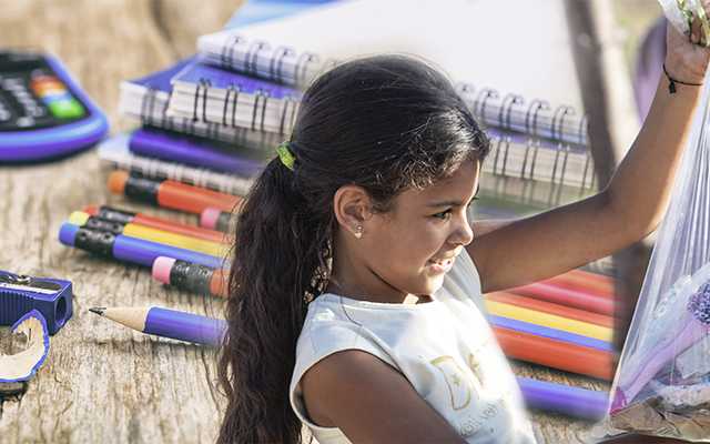 Menina segurando um kit escolar embrulhado. Ao fundo, material escolar como cadernos, lápis e borracha.