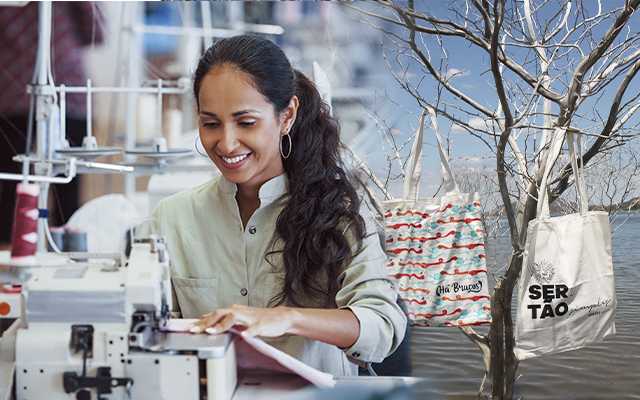 Mulher costurando em uma máquina de costura. Ao fundo, uma árvore sem folhas em frente a um rio, com sacolas do Gretas penduradas em seus galhos.