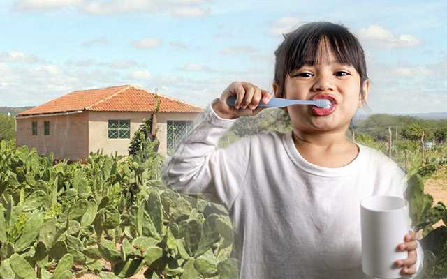 Menina escovando os dentes. No fundo, uma casa simples em meio a uma plantação de palma.